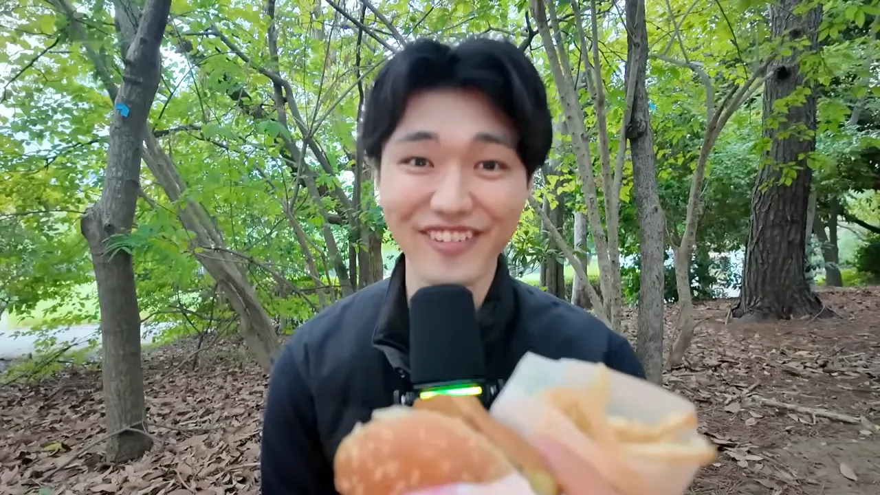 昼の公園で、ハンバーガーとポテトを画面の手前に掲げて笑っている場面